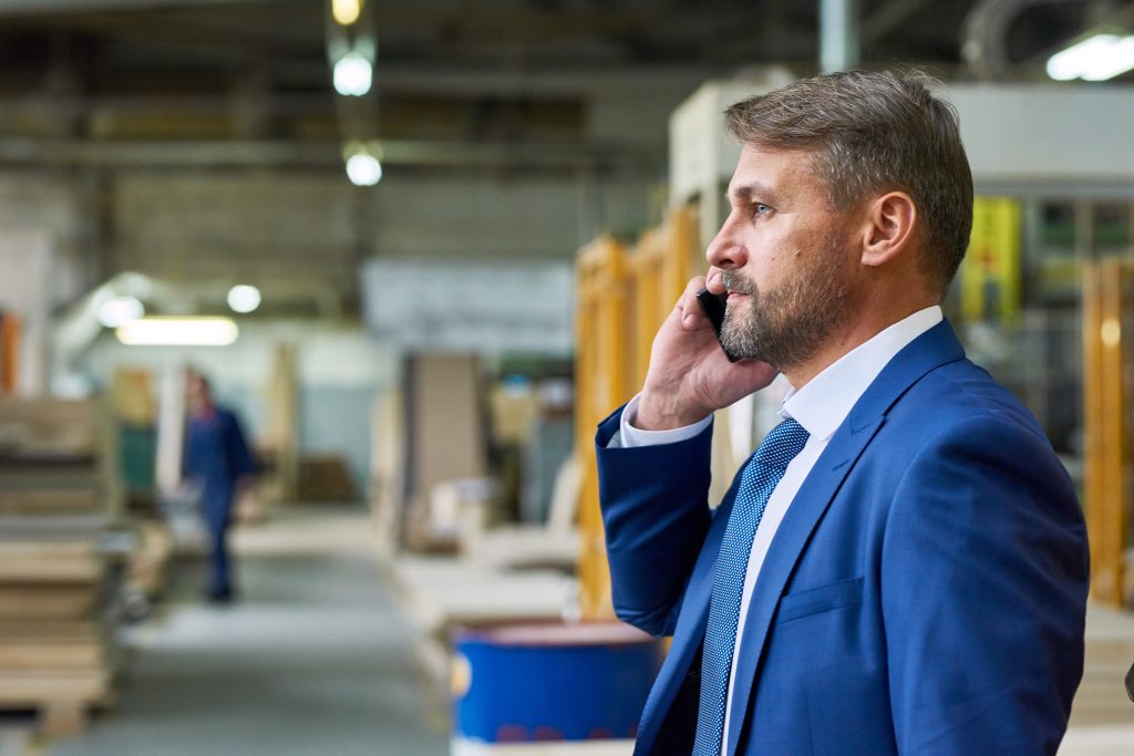 Waist up side view of handsome mature businessman speaking by phone in factory workshop with workers in background, copy space